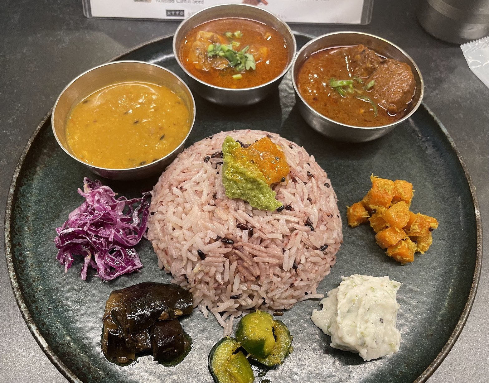 黒いプレートに紫米のライスを中心に、3つのステンレスボウルにカレー、周囲に副菜6〜7種が配置されたプレート全景