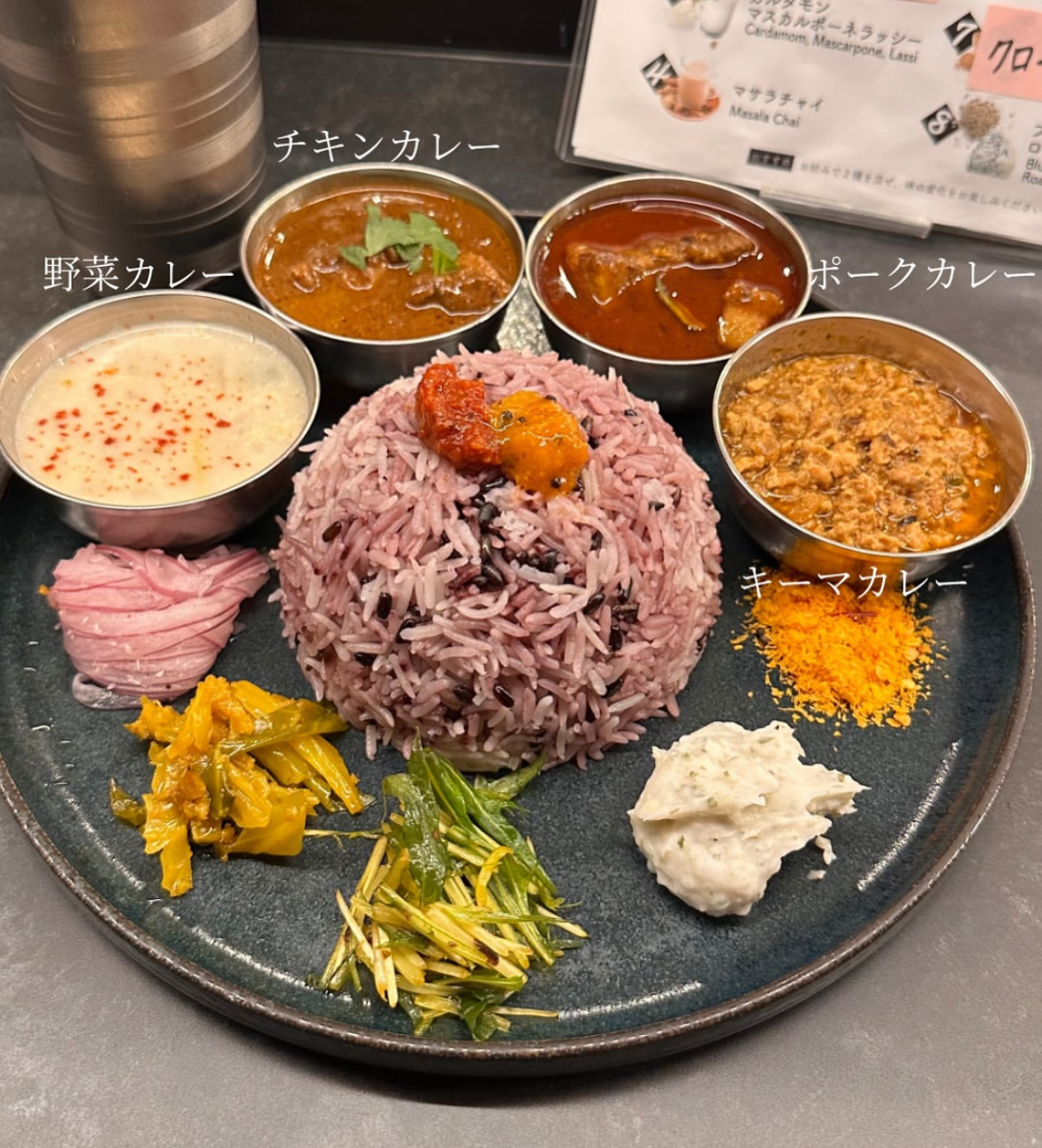 ステンレスボウルに「チキンカレー」「野菜カレー」「ポークカレー」「キーマカレー」のラベルが付き、紫米ライスと副菜が並ぶプレート
