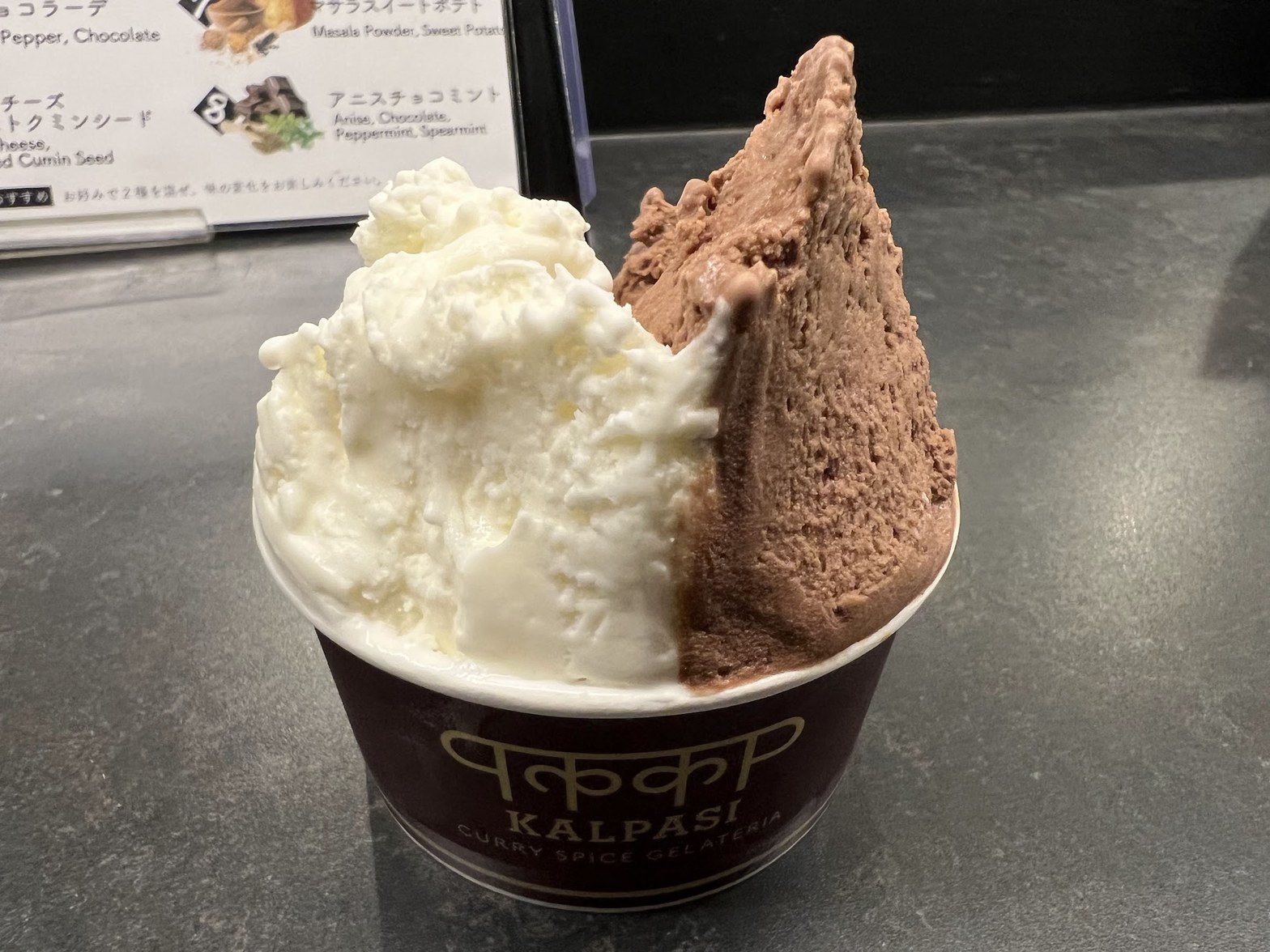 KALPASIのロゴ入り茶色い紙カップに、白いジェラートとチョコレート色のジェラートが2種盛りで提供されている